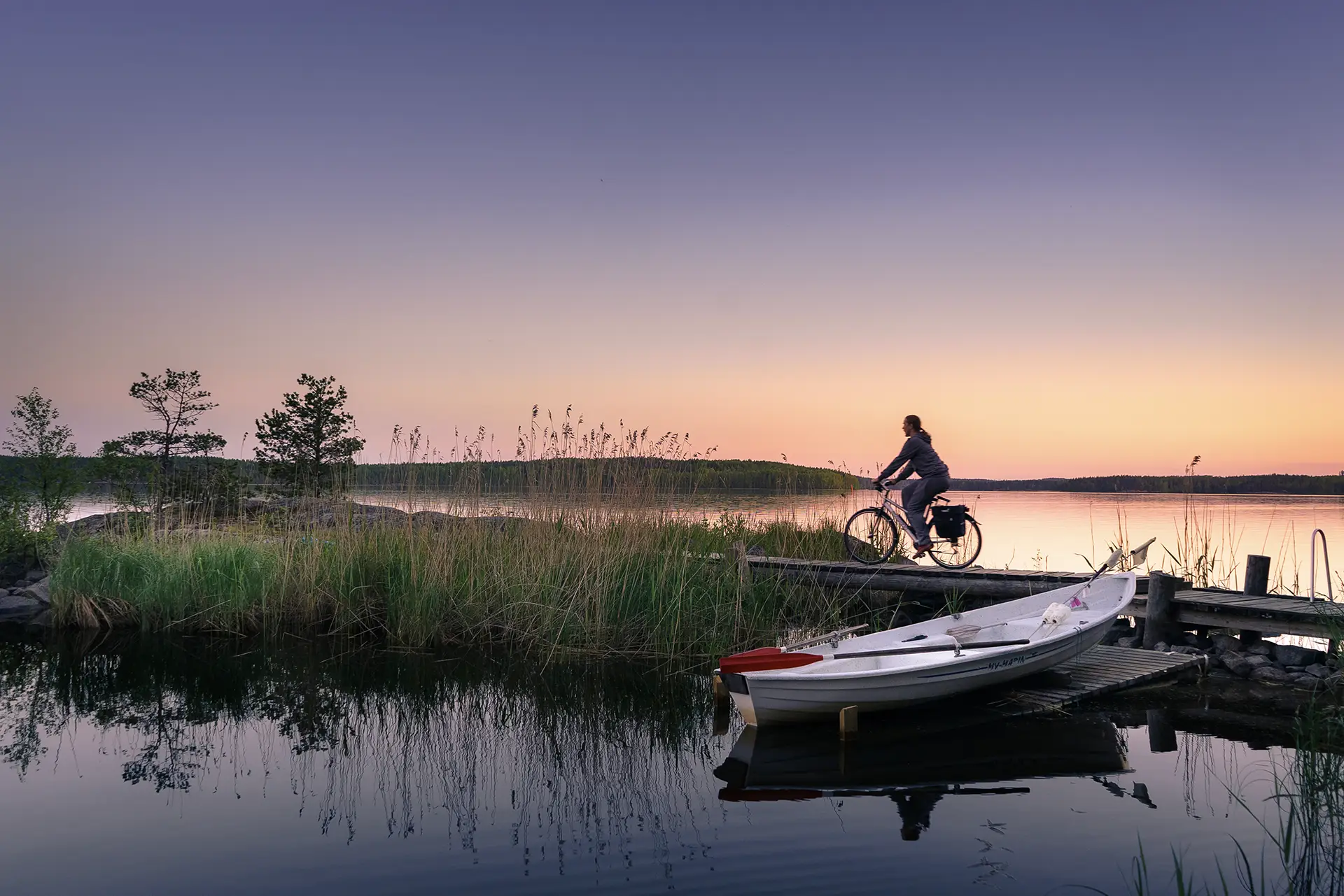 Nainen ajaa pyörällä kauniissa maisemassa Saimaalla veden äärellä. Edustalla soutuvene
