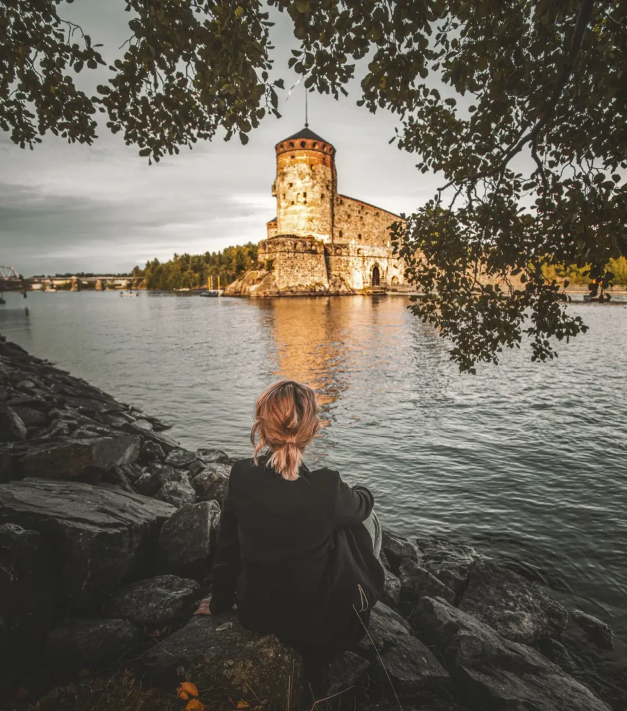 Nainen istuu rannalla ja ihailee näkymää Olavinlinnaan