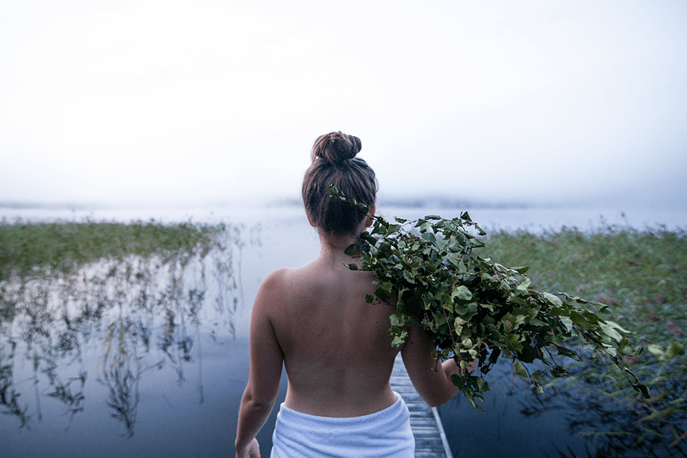 nainen kävelee poispäin kamerasta kohti utuista järveä kantaen saunavastaa olallaan
